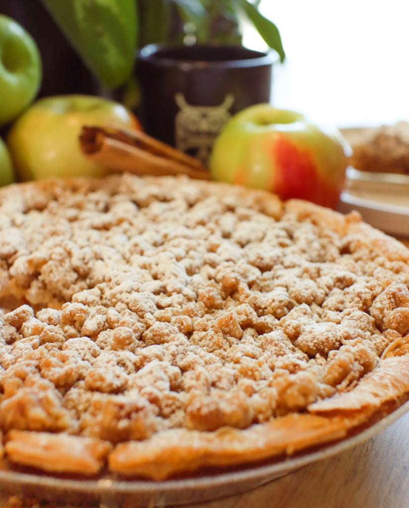 close up image of second state coffee's holiday apple pie, dusted with powdered sugar. apples and cinnamon sticks blurred in background
