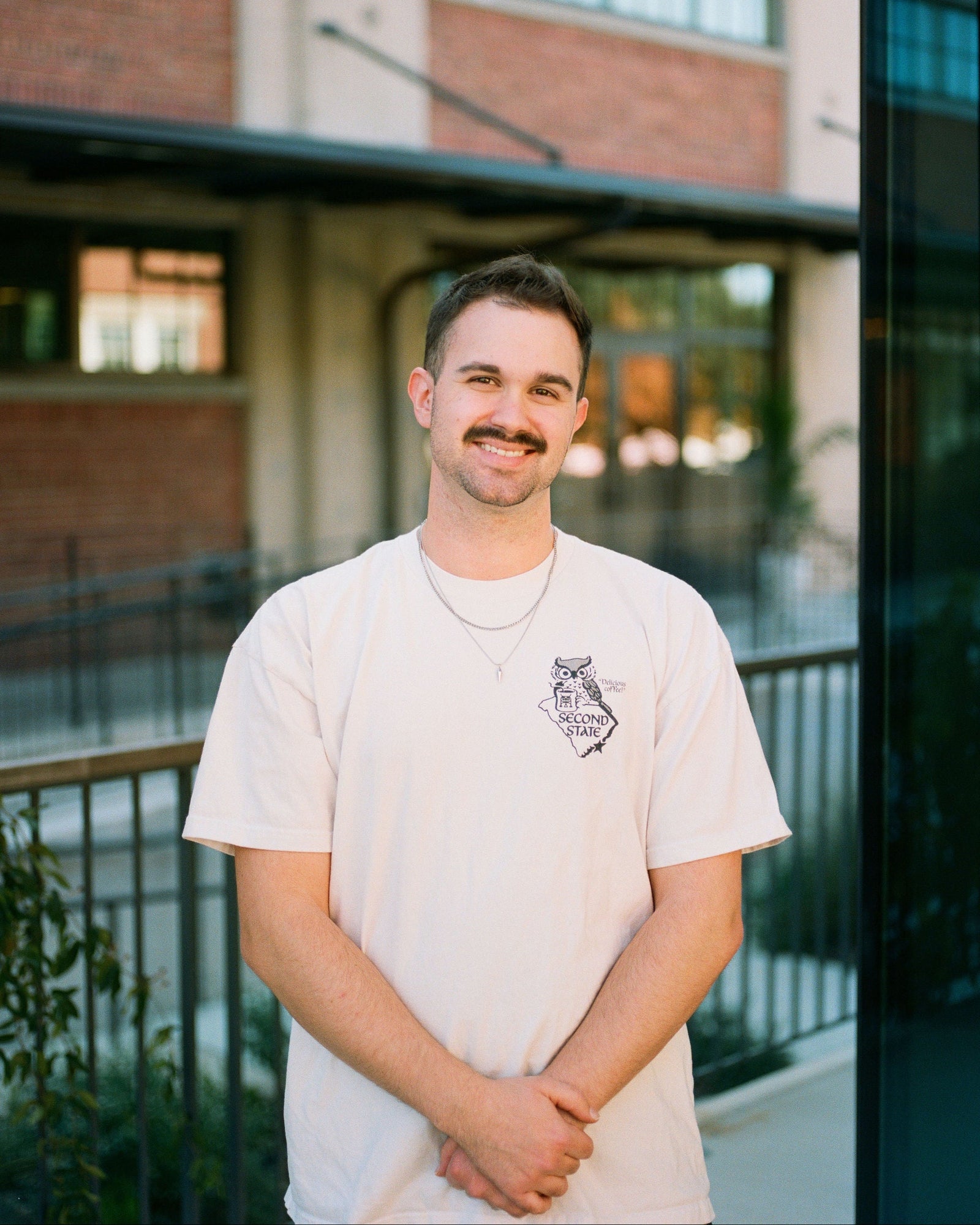 Man wearing a light-colored t-shirt with a second state coffee logo, standing outdoors with a building in the background.