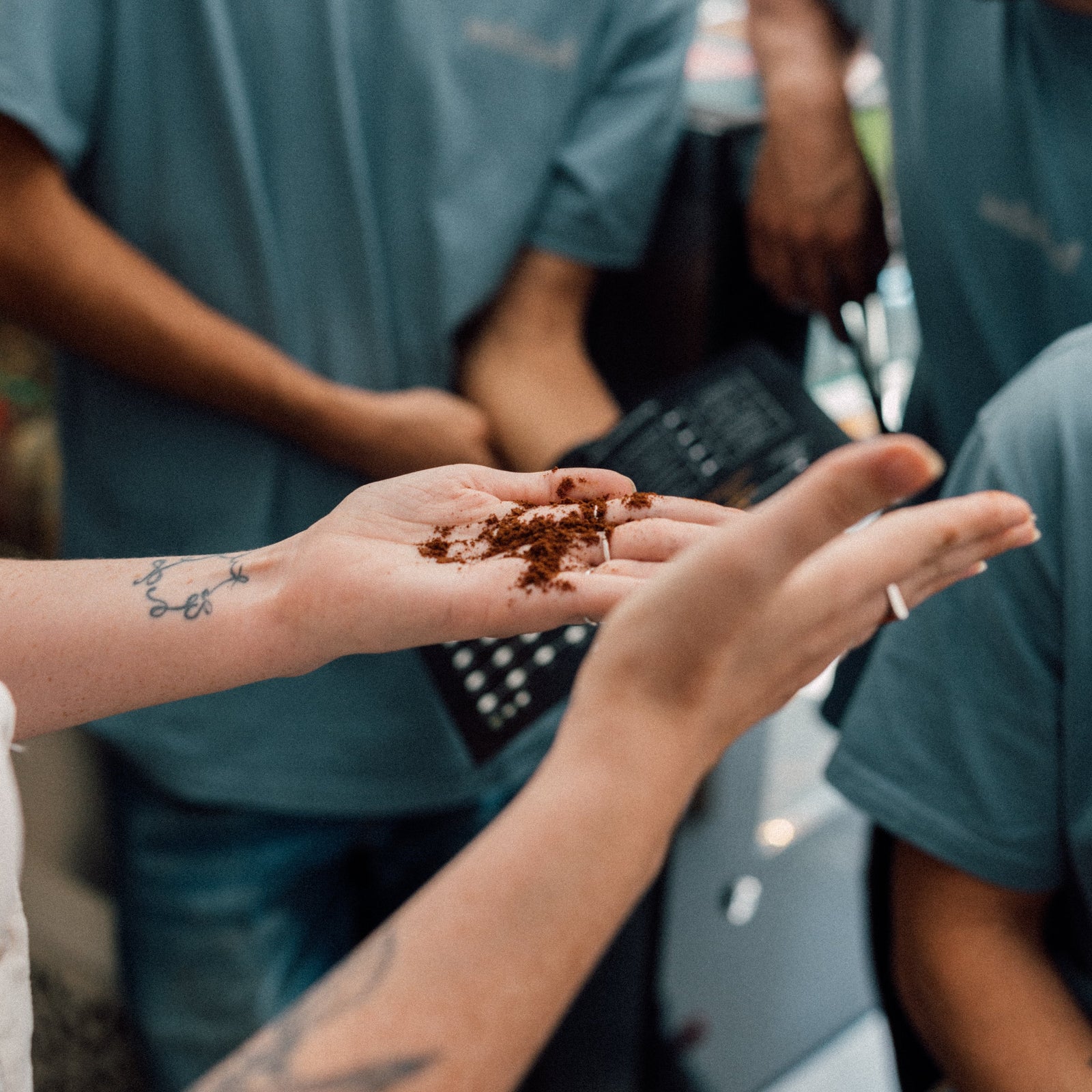 Person holding coffee grounds in their hand with two individuals in teal shirts in the background. Demonstrates specialty coffee barista classroom training coffee professionals in espresso