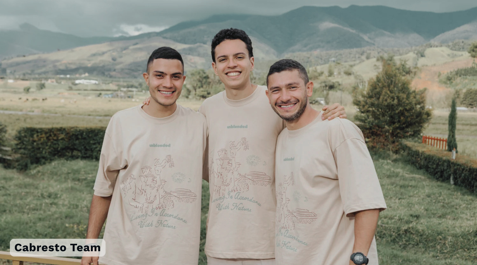 Three coffee producers standing outdoors in front of mountains, wearing matching beige Unblended graphic t-shirts. The t-shirt features a coffee farm illustration. The banner displays text in the bottom left corner reading 'Cabresto Team'.