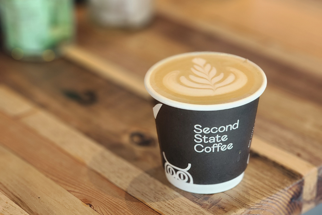 Coffee cup with latte art on a wooden surface, featuring 'Second State Coffee'.