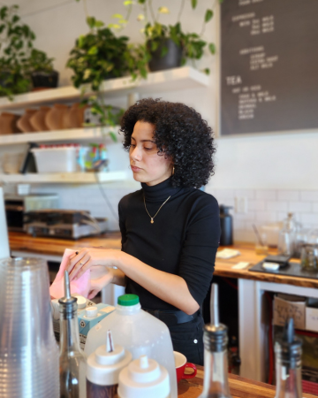 Person working behind a counter in a coffee shop making a coffee, syrup bottles and cups in the foreground, a menu hanging on the wall in background
