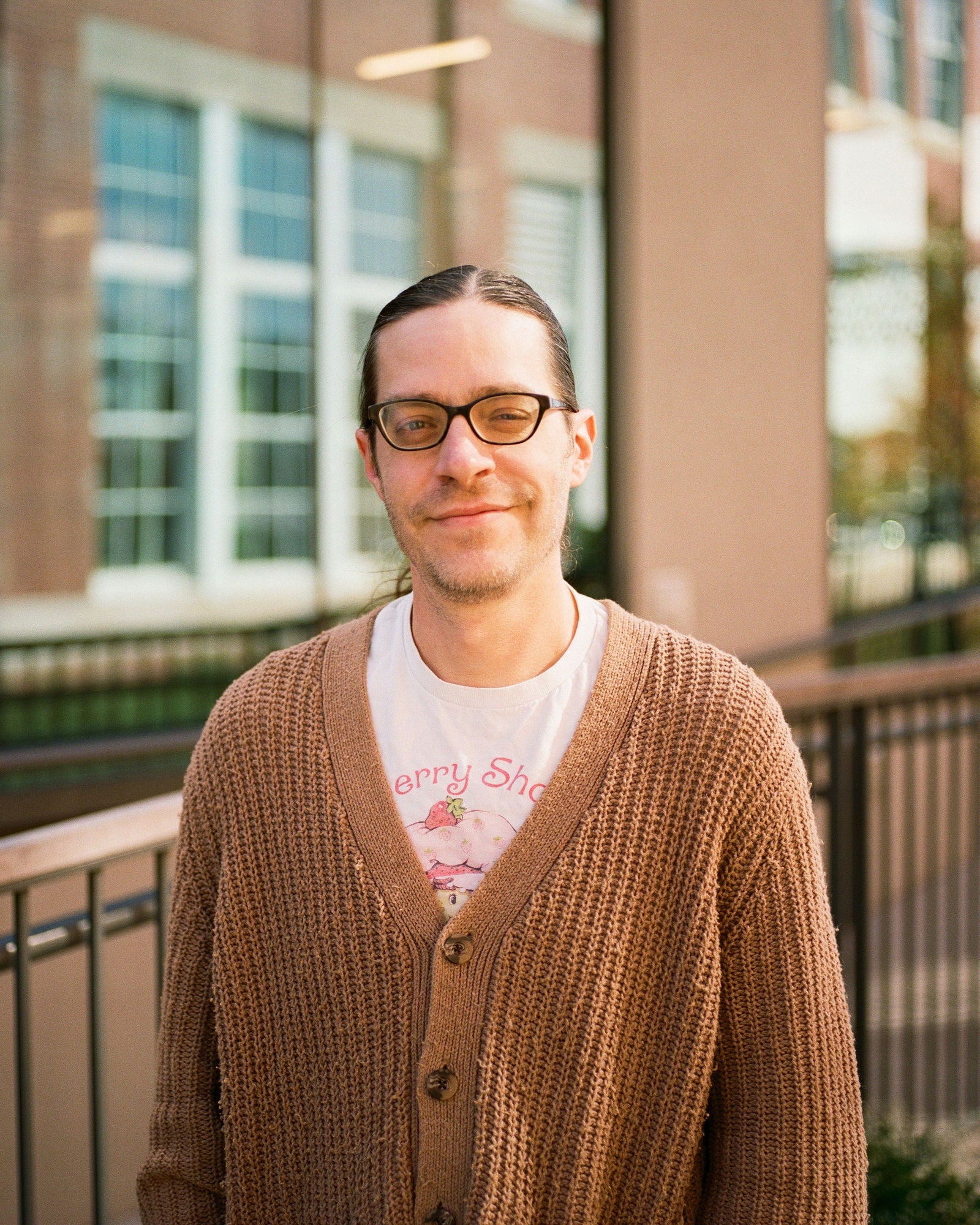 Person wearing a brown cardigan over a white shirt with text, standing outdoors.