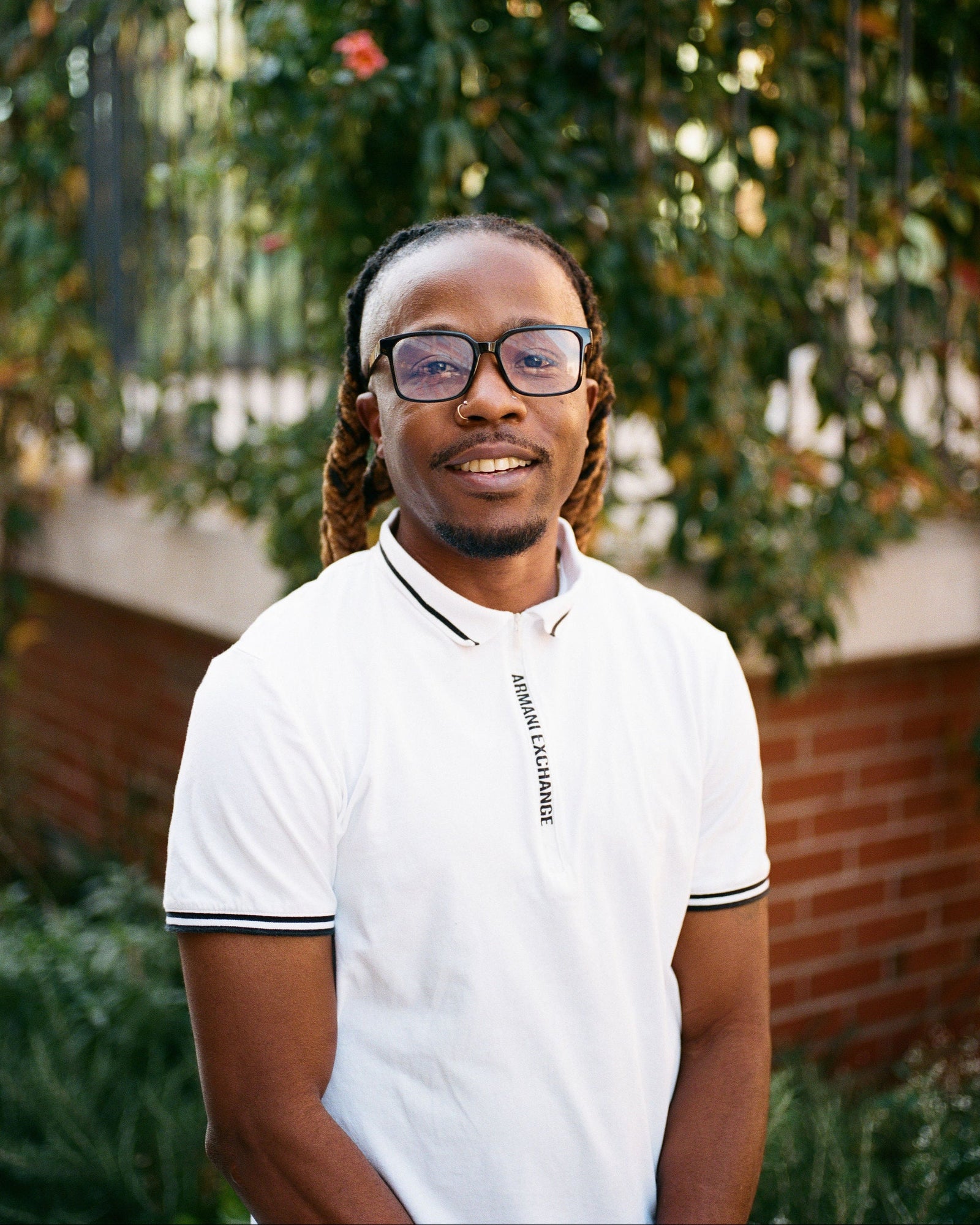 Person wearing a white polo shirt with text, standing outdoors with greenery in the background